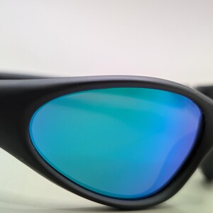 May include: Close-up of black sunglasses with blue and green mirrored lenses. The sunglasses have a sleek, modern design with a curved frame and a small silver emblem on the side. The lenses reflect a gradient of blue and green.