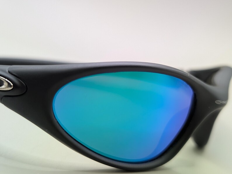 May include: Close-up of black sunglasses with blue and green mirrored lenses. The sunglasses have a sleek, modern design with a curved frame and a small silver emblem on the side. The lenses reflect a gradient of blue and green.