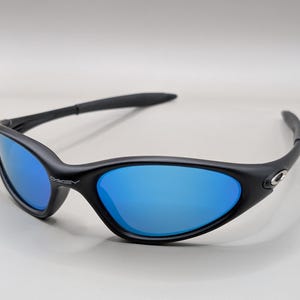 K&ouml;nnte beinhalten: Schwarze Sonnenbrille mit blau verspiegelten Gl&auml;sern. Die Sonnenbrille hat ein geschwungenes, umlaufendes Design mit einem schwarzen Rahmen. Der Markenname "Oakley" ist auf dem Rahmen sichtbar.