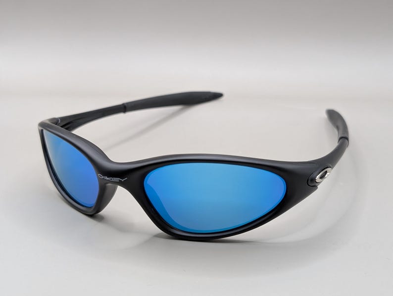 K&ouml;nnte beinhalten: Schwarze Sonnenbrille mit blau verspiegelten Gl&auml;sern. Die Sonnenbrille hat ein geschwungenes, umlaufendes Design mit einem schwarzen Rahmen. Der Markenname "Oakley" ist auf dem Rahmen sichtbar.
