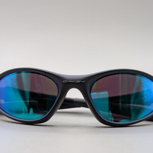 May include: Black oval-shaped sunglasses with blue and purple reflective lenses. The sunglasses have a matte finish and the brand name "Oakley" is visible on the frame. The arms are folded.