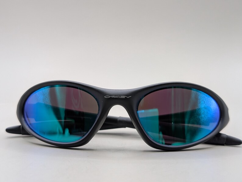 May include: Black oval-shaped sunglasses with blue and purple reflective lenses. The sunglasses have a matte finish and the brand name "Oakley" is visible on the frame. The arms are folded.