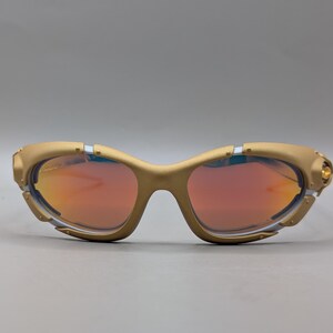 May include: Gold-colored sunglasses with red-tinted lenses. The sunglasses have a curved frame with a unique design, featuring small silver and gold accents. The lenses are a vibrant red color, and the overall style is sporty and modern.