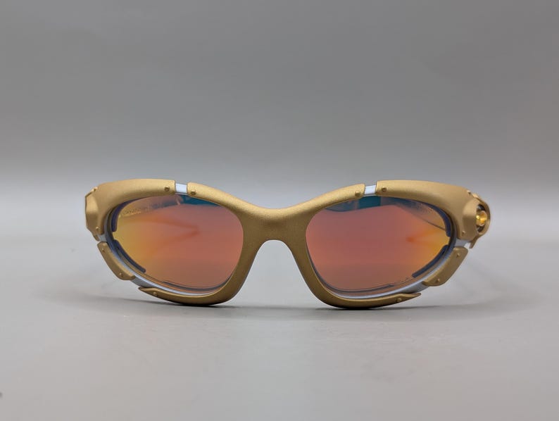 May include: Gold-colored sunglasses with red-tinted lenses. The sunglasses have a curved frame with a unique design, featuring small silver and gold accents. The lenses are a vibrant red color, and the overall style is sporty and modern.