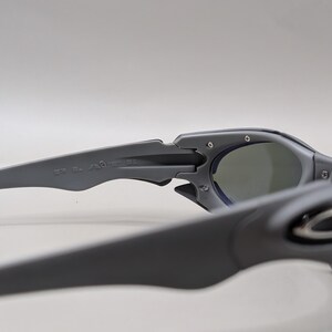 May include: Close-up of gray sunglasses with dark lenses. The sunglasses have a sleek, modern design with a matte finish. The arms of the sunglasses are thick and feature the brand name. The lenses are a dark gray color.