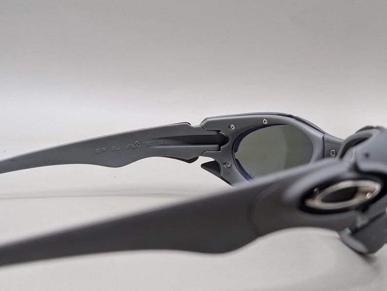 May include: Close-up of gray sunglasses with dark lenses. The sunglasses have a sleek, modern design with a matte finish. The arms of the sunglasses are thick and feature the brand name. The lenses are a dark gray color.