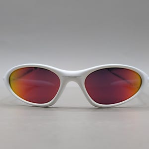 May include: White oval sunglasses with a red-orange gradient lens. The sunglasses have a white frame and the brand name "Oakley" is visible on the frame. The lenses have a reflective quality.
