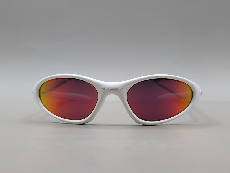 May include: White oval sunglasses with a red-orange gradient lens. The sunglasses have a white frame and the brand name "Oakley" is visible on the frame. The lenses have a reflective quality.