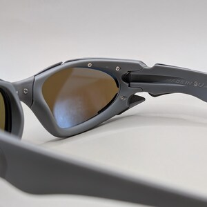 K&ouml;nnte beinhalten: Graue Sonnenbrille mit braun get&ouml;nten Gl&auml;sern. Der matte graue Rahmen tr&auml;gt den Schriftzug "MADE IN U.S.A." auf dem B&uuml;gel. Das Design weist eine markante Scharnier- und B&uuml;gelform auf, geeignet f&uuml;r Sport oder Outdoor-Aktivit&auml;ten.