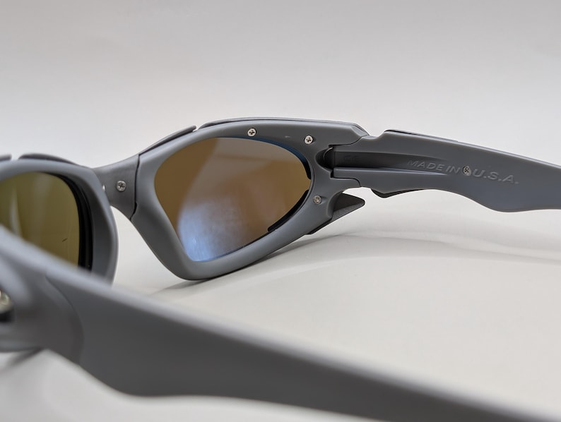 K&ouml;nnte beinhalten: Graue Sonnenbrille mit braun get&ouml;nten Gl&auml;sern. Der matte graue Rahmen tr&auml;gt den Schriftzug "MADE IN U.S.A." auf dem B&uuml;gel. Das Design weist eine markante Scharnier- und B&uuml;gelform auf, geeignet f&uuml;r Sport oder Outdoor-Aktivit&auml;ten.