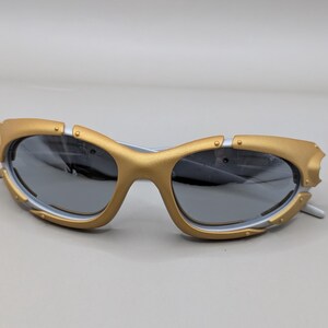 May include: Gold and white sunglasses with gray lenses. The sunglasses have a unique design with gold accents around the lenses and a white frame. The lenses are a dark gray color, providing a sleek look.