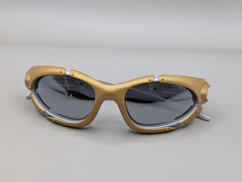 May include: Gold and white sunglasses with gray lenses. The sunglasses have a unique design with gold accents around the lenses and a white frame. The lenses are a dark gray color, providing a sleek look.