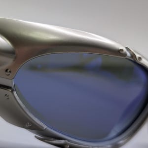 May include: Close-up of silver sunglasses with blue lenses. The sunglasses have a modern design with visible rivets and a curved frame. The lenses reflect the surrounding environment. The frame is made of metal.