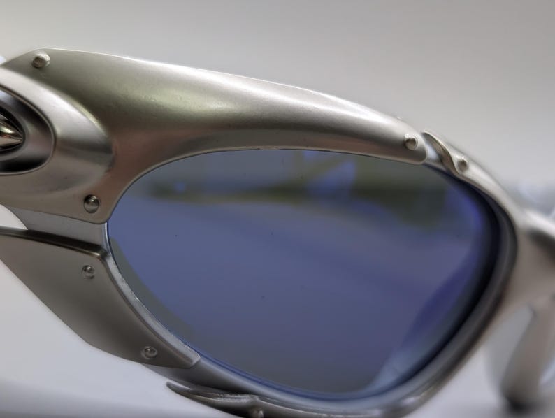 May include: Close-up of silver sunglasses with blue lenses. The sunglasses have a modern design with visible rivets and a curved frame. The lenses reflect the surrounding environment. The frame is made of metal.