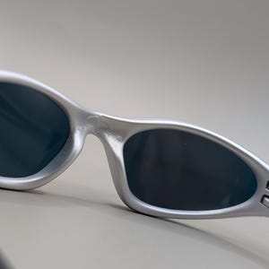 May include: Silver-framed sunglasses with dark lenses. The sunglasses have a sleek, curved design with the word "MINUTE" printed on the frame. The temples have a unique, angular shape.