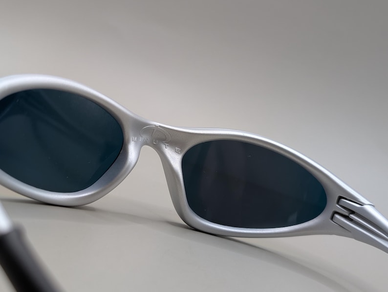 May include: Silver-framed sunglasses with dark lenses. The sunglasses have a sleek, curved design with the word "MINUTE" printed on the frame. The temples have a unique, angular shape.