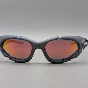 Puede incluir: Gafas de sol deportivas gris oscuro con lentes naranja-rojo. Las gafas tienen un dise&ntilde;o envolvente con una montura gruesa y un peque&ntilde;o acento plateado en el lateral. Las lentes tienen un ligero tinte azul en los bordes.