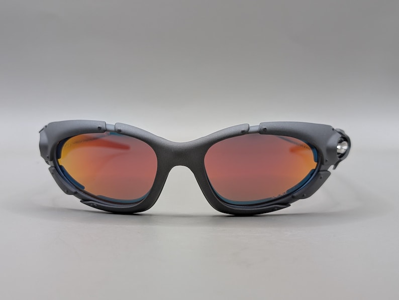 Puede incluir: Gafas de sol deportivas gris oscuro con lentes naranja-rojo. Las gafas tienen un dise&ntilde;o envolvente con una montura gruesa y un peque&ntilde;o acento plateado en el lateral. Las lentes tienen un ligero tinte azul en los bordes.