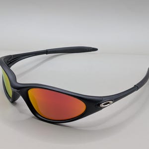 Pode incluir: &Oacute;culos de sol pretos com lentes vermelho-alaranjadas. Os &oacute;culos de sol t&ecirc;m um design elegante e curvo com acabamento fosco. As lentes refletem um gradiente de cores, e a arma&ccedil;&atilde;o apresenta um pequeno detalhe prateado perto da haste.