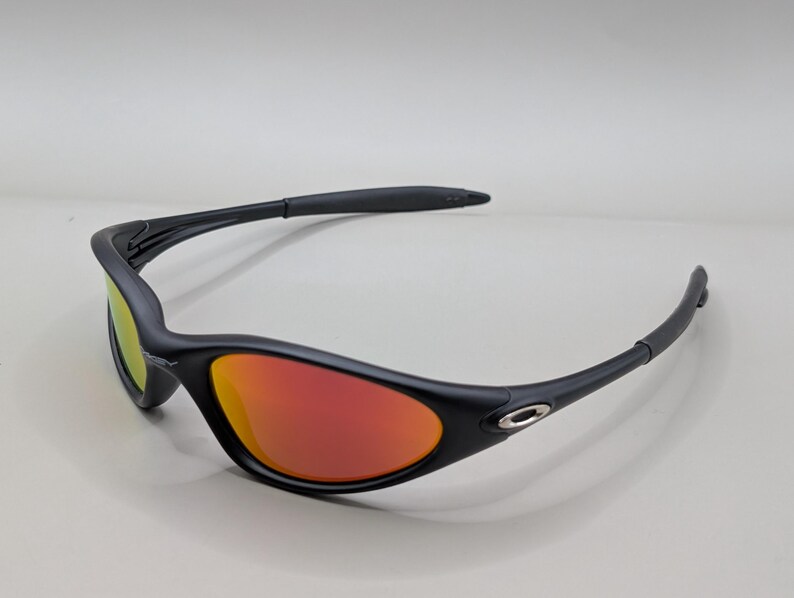 Pode incluir: &Oacute;culos de sol pretos com lentes vermelho-alaranjadas. Os &oacute;culos de sol t&ecirc;m um design elegante e curvo com acabamento fosco. As lentes refletem um gradiente de cores, e a arma&ccedil;&atilde;o apresenta um pequeno detalhe prateado perto da haste.