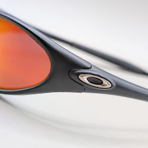 Pode incluir: Close-up de &oacute;culos de sol pretos com lentes gradientes laranja e amarelo. Os &oacute;culos de sol t&ecirc;m um design elegante com um detalhe prateado na haste. As lentes s&atilde;o curvas e refletem a luz.
