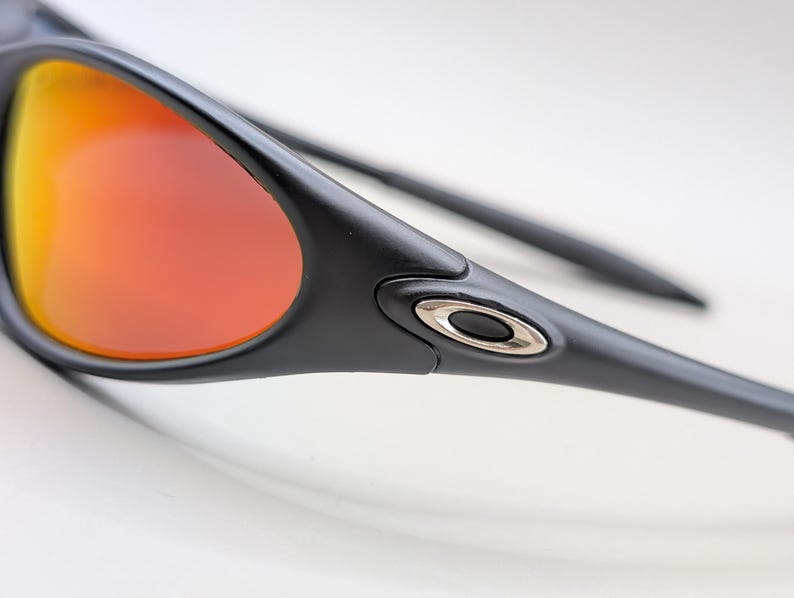 Pode incluir: Close-up de &oacute;culos de sol pretos com lentes gradientes laranja e amarelo. Os &oacute;culos de sol t&ecirc;m um design elegante com um detalhe prateado na haste. As lentes s&atilde;o curvas e refletem a luz.