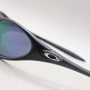 May include: Close-up of black sunglasses with a sleek, modern design. The lenses have a reflective, multi-colored coating, and the frame features a silver emblem. The sunglasses are angled, showcasing their streamlined shape and sporty aesthetic.