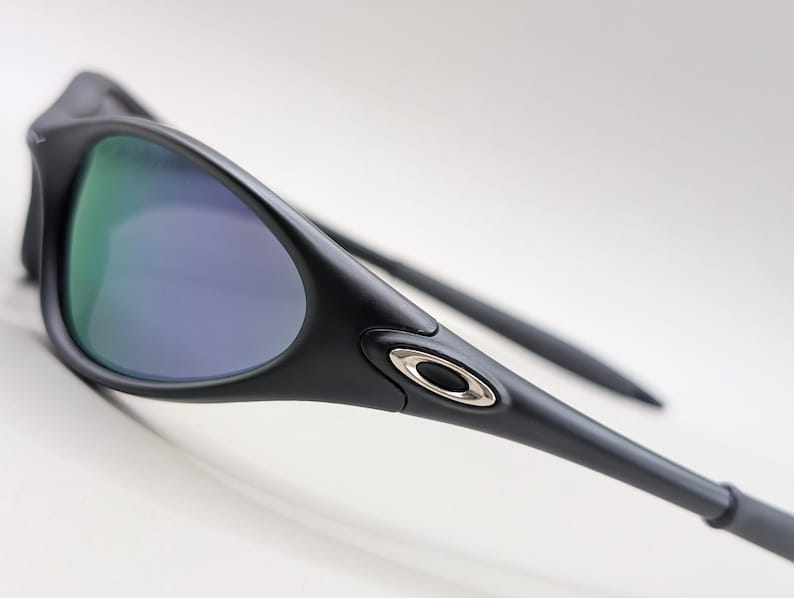 May include: Close-up of black sunglasses with a sleek, modern design. The lenses have a reflective, multi-colored coating, and the frame features a silver emblem. The sunglasses are angled, showcasing their streamlined shape and sporty aesthetic.
