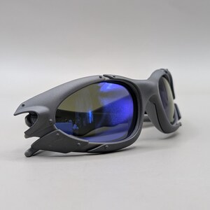 May include: Matte gray sunglasses with blue lenses. The sunglasses have a unique, angular design with a futuristic aesthetic. The lenses reflect a blue hue, and the frames have a textured finish.