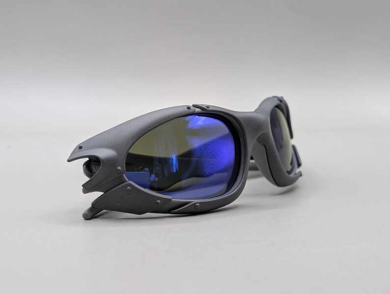 May include: Matte gray sunglasses with blue lenses. The sunglasses have a unique, angular design with a futuristic aesthetic. The lenses reflect a blue hue, and the frames have a textured finish.