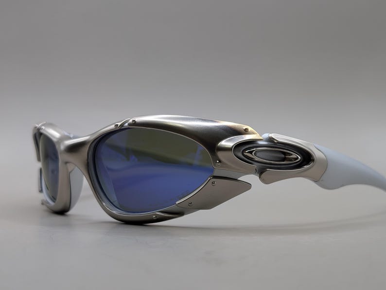 May include: Silver and white sunglasses with blue lenses. The sunglasses feature a futuristic design with a metallic frame and white arms. The lenses are a vibrant blue, and the overall style is sporty and modern.