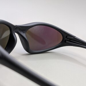 May include: Black sunglasses with a curved frame and tinted lenses. The lenses have a reflective, purple hue. The arms of the sunglasses are sleek and feature a hinge design. The brand name is visible on the frame.