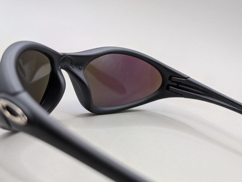 May include: Black sunglasses with a curved frame and tinted lenses. The lenses have a reflective, purple hue. The arms of the sunglasses are sleek and feature a hinge design. The brand name is visible on the frame.