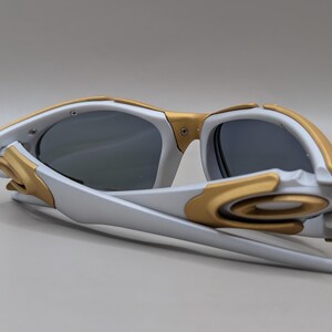 K&ouml;nnte beinhalten: Wei&szlig;-goldene Sonnenbrille mit dunklen Gl&auml;sern. Die Sonnenbrille hat ein modernes, eckiges Design mit einem wei&szlig;en Rahmen und goldenen Details um die Gl&auml;ser und B&uuml;gel. Die Gl&auml;ser sind dunkelgrau.