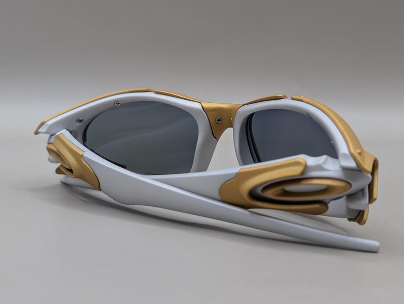K&ouml;nnte beinhalten: Wei&szlig;-goldene Sonnenbrille mit dunklen Gl&auml;sern. Die Sonnenbrille hat ein modernes, eckiges Design mit einem wei&szlig;en Rahmen und goldenen Details um die Gl&auml;ser und B&uuml;gel. Die Gl&auml;ser sind dunkelgrau.