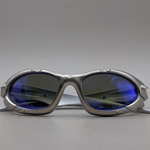 May include: Silver sunglasses with blue-tinted lenses. The sunglasses have a futuristic design with a curved frame and a bridge. The lenses are oval-shaped and reflect light. The temples are thin and extend back.