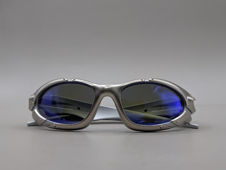 May include: Silver sunglasses with blue-tinted lenses. The sunglasses have a futuristic design with a curved frame and a bridge. The lenses are oval-shaped and reflect light. The temples are thin and extend back.
