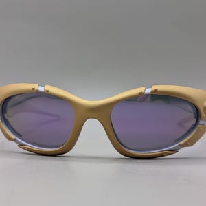 K&ouml;nnte beinhalten: Goldfarbene Sonnenbrille mit violetten Gl&auml;sern. Die Fassungen haben ein einzigartiges, futuristisches Design mit silbernen Akzenten und sichtbaren Scharnieren. Die Gl&auml;ser sind tiefviolett und bilden einen Kontrast zum goldenen Rahmen.