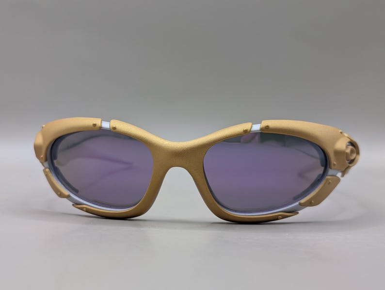 K&ouml;nnte beinhalten: Goldfarbene Sonnenbrille mit violetten Gl&auml;sern. Die Fassungen haben ein einzigartiges, futuristisches Design mit silbernen Akzenten und sichtbaren Scharnieren. Die Gl&auml;ser sind tiefviolett und bilden einen Kontrast zum goldenen Rahmen.