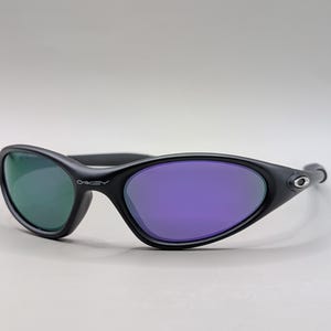 May include: Black Oakley sunglasses with a curved frame and oval lenses. One lens is green, the other is purple. The Oakley logo is visible on the frame.