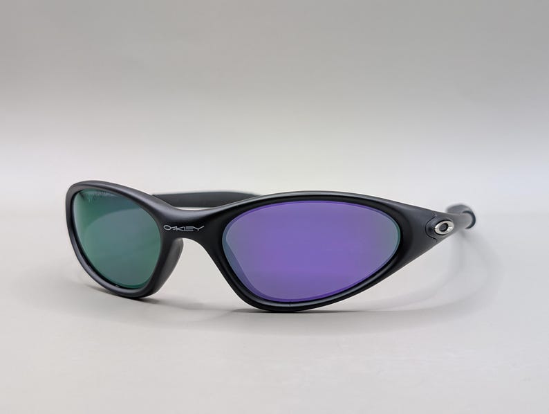 May include: Black Oakley sunglasses with a curved frame and oval lenses. One lens is green, the other is purple. The Oakley logo is visible on the frame.