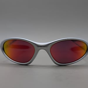 May include: Silver-framed sunglasses with red and orange gradient lenses. The sunglasses have a curved, wraparound design and the brand name is visible on the frame. The lenses transition from red to orange.
