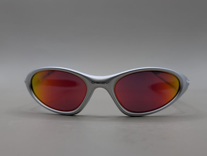 May include: Silver-framed sunglasses with red and orange gradient lenses. The sunglasses have a curved, wraparound design and the brand name is visible on the frame. The lenses transition from red to orange.