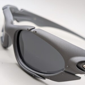 May include: Close-up of gray sunglasses with dark lenses. The sunglasses have a matte finish and a sporty design. The frame features a unique, angular shape with visible hinges and a silver accent near the temple.