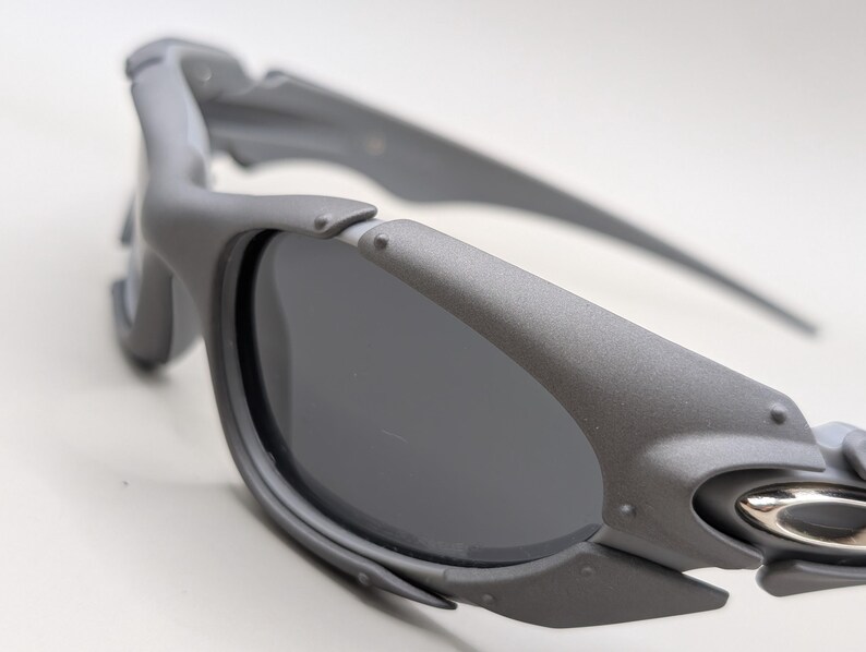 May include: Close-up of gray sunglasses with dark lenses. The sunglasses have a matte finish and a sporty design. The frame features a unique, angular shape with visible hinges and a silver accent near the temple.
