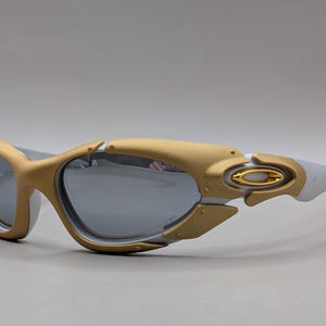 May include: Gold and white sunglasses with gray lenses. The sunglasses have a gold frame around the lenses and a white frame on the arms. The lenses are reflective. The sunglasses are a sporty style.