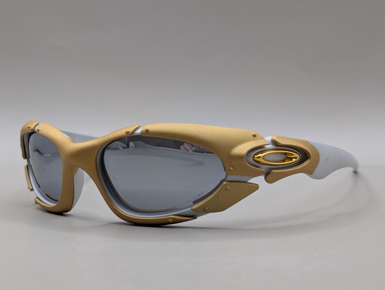 May include: Gold and white sunglasses with gray lenses. The sunglasses have a gold frame around the lenses and a white frame on the arms. The lenses are reflective. The sunglasses are a sporty style.