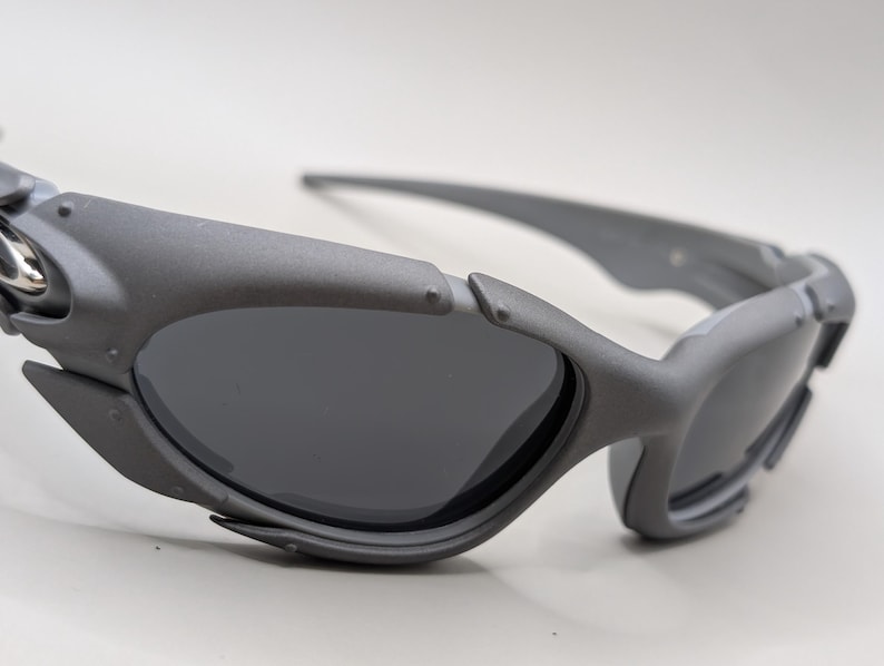 May include: Close-up of gray sunglasses with dark lenses. The sunglasses have a futuristic design with a matte finish. The arms and frame are thick and angular, with a small silver accent near the hinge.
