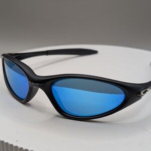 K&ouml;nnte beinhalten: Schwarze Sonnenbrille mit blau verspiegelten Gl&auml;sern. Die Sonnenbrille hat einen geschwungenen Rahmen und B&uuml;gel. Das Oakley-Logo ist an der Seite des Rahmens sichtbar.