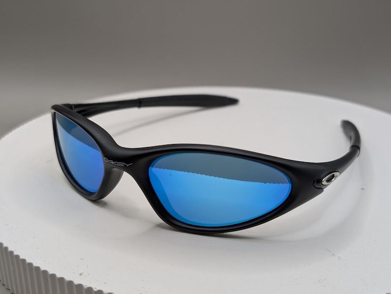 K&ouml;nnte beinhalten: Schwarze Sonnenbrille mit blau verspiegelten Gl&auml;sern. Die Sonnenbrille hat einen geschwungenen Rahmen und B&uuml;gel. Das Oakley-Logo ist an der Seite des Rahmens sichtbar.